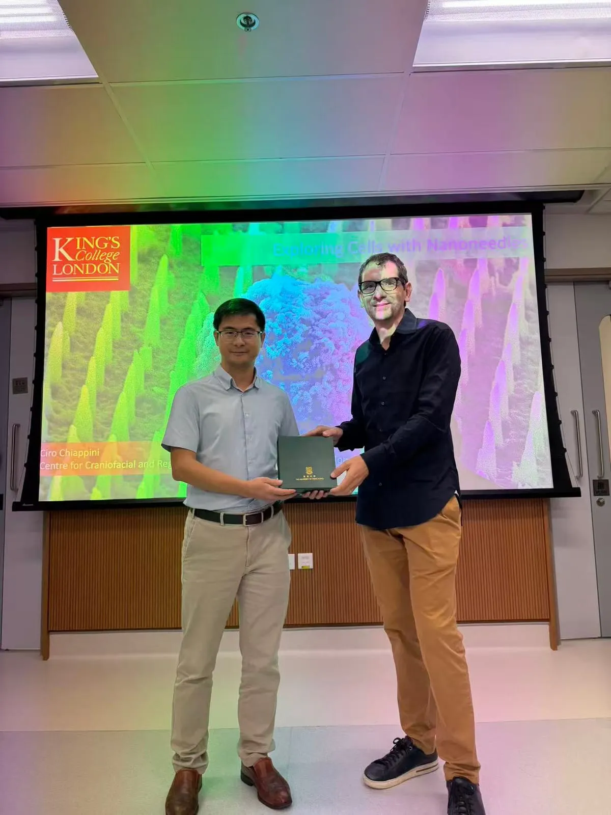 Ciro Chiappini receiving a gift from a host after his HKU talk, with the title slide ‘Exploring Cells with Nanoneedles’ on the screen behind them