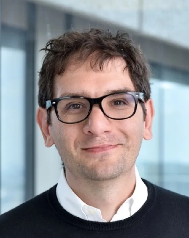 Portrait of Ciro Chiappini, smiling, wearing glasses and a dark jumper over a white shirt.