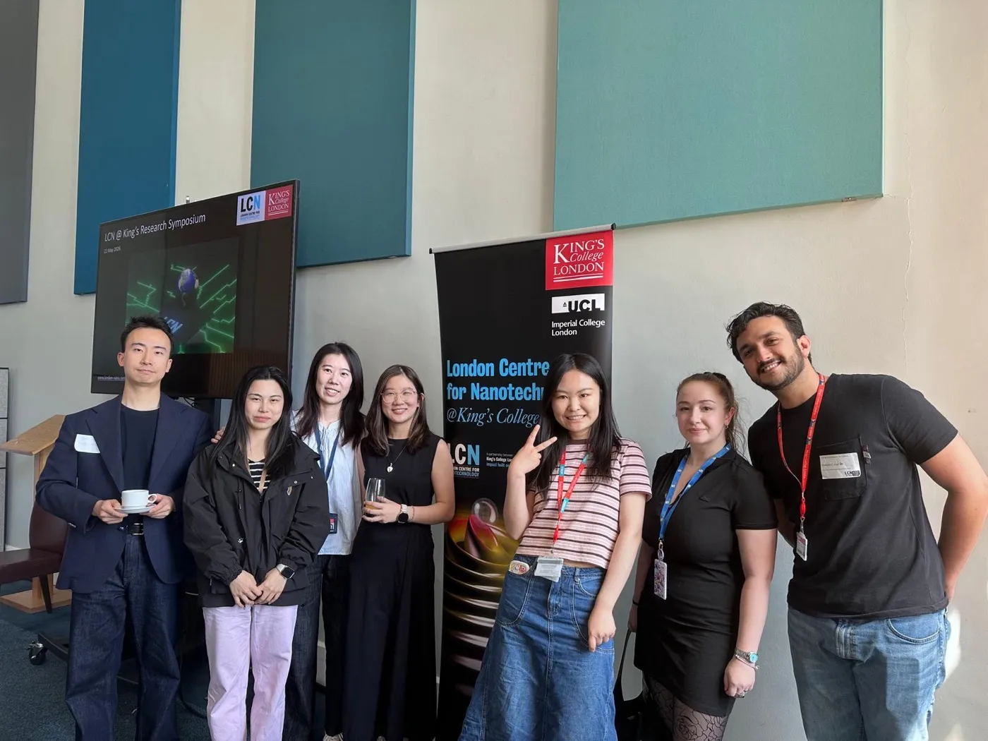 Seven lab members standing beside a London Centre for Nanotechnology banner at a research symposium.